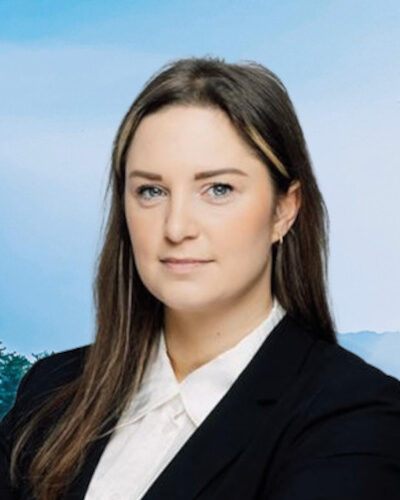 Eine Frau mit dunklen Haaren und formeller Kleidung steht vor einem blauen Hintergrund.